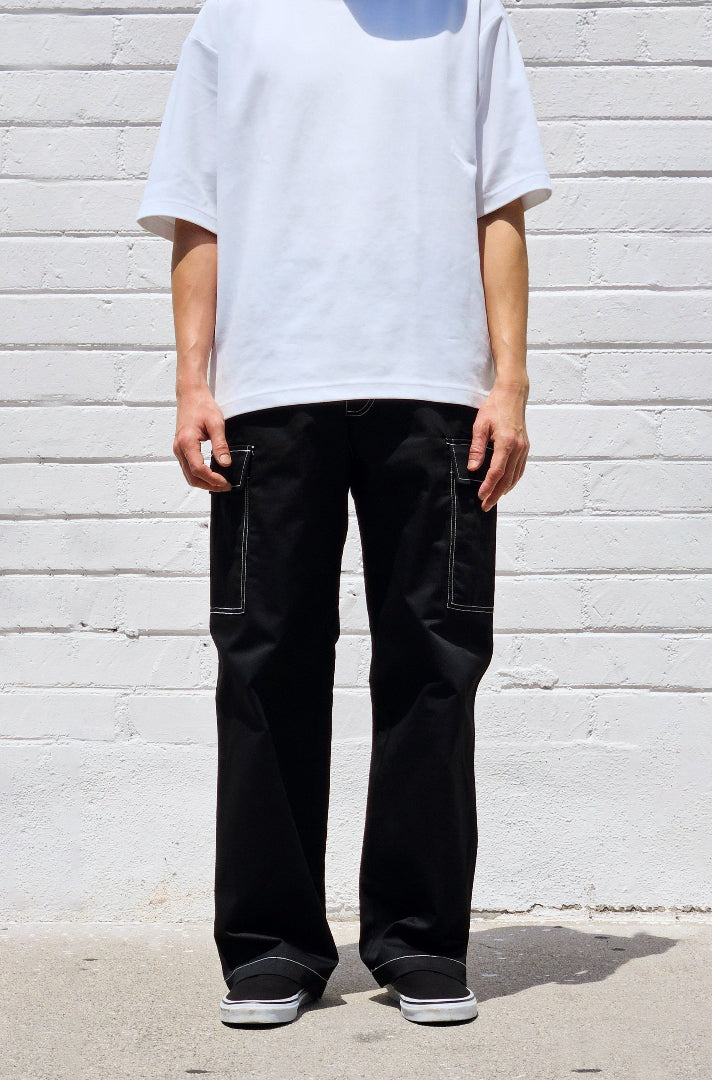 Person wearing a white t-shirt and black pants against a light gray brick wall.