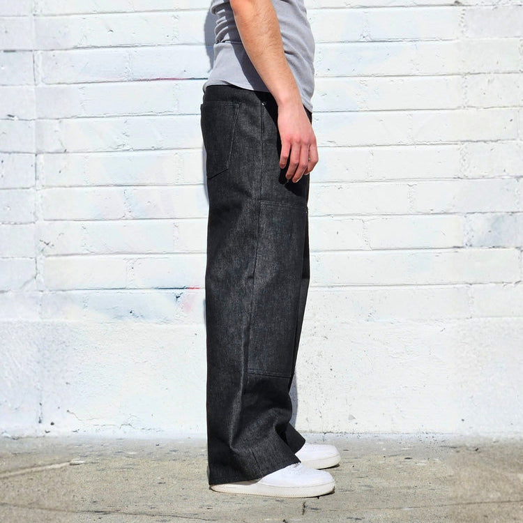 Person wearing a gray tank top and dark jeans standing against a light brick wall.