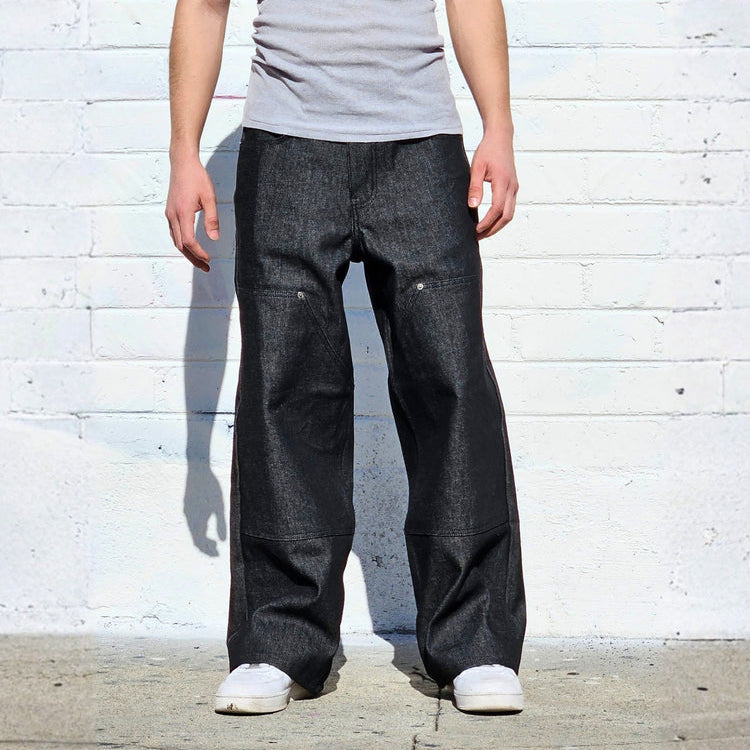 Person wearing a gray tank top and dark jeans standing against a white brick wall.